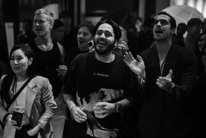 A black-and-white photo shows a bearded man wearing a 'PHOENIX' t-shirt, singing alongside a cheering crowd with raised hands.