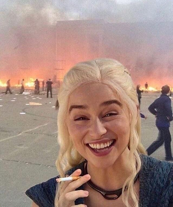A smiling blonde woman holds a cigarette in front of a raging fire and chaotic background, with the caption "Me thinking about how to turn my last $50 into $1B" humorously suggesting she's carelessly planning risky investments while everything burns around her. The juxtaposition of her blissful smile against the apocalyptic backdrop satirizes reckless optimism and YOLO financial decision-making.