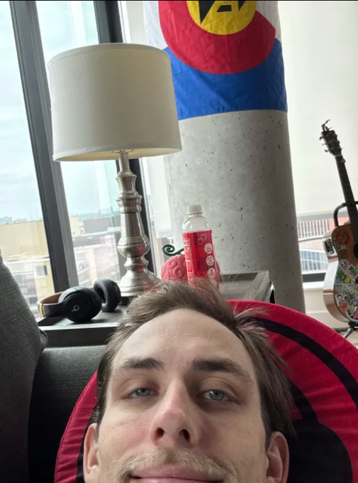 A man takes a selfie in a red chair looking exhausted, with a lamp, headphones, and a guitar visible behind him against a backdrop of windows. The "gm @Stake" caption references the crypto gambling platform Stake, while his tired appearance and mention of "5 hours of sleep" set the stage for a day of risky betting.