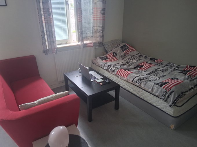 A compact 15m² bedroom featuring a red armchair, black coffee table with a laptop, and a single bed with American flag-patterned bedding, embodying a focused "grind" workspace setup. The minimalistic layout reflects the disciplined, productive energy of the caption's message about locking in and making noise.