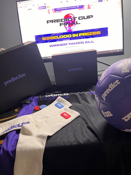 A promotional setup for "Predict Cup Final" featuring the predict.fun brand on boxes, a jersey with YES/NO voting buttons, and a branded soccer ball, all displayed in front of a screen advertising a "$260,000 IN PRIZES" contest with the tagline "WINNER TAKES ALL"—visually representing the project's prediction-based gaming platform mentioned in the tweet.