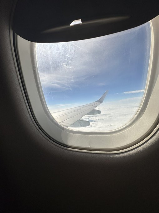 Looking out from an airplane window, the wing stretches above a blanket of clouds under a clear blue sky—a moment of calm transit that reflects the "web3 idol's" journey between destinations.