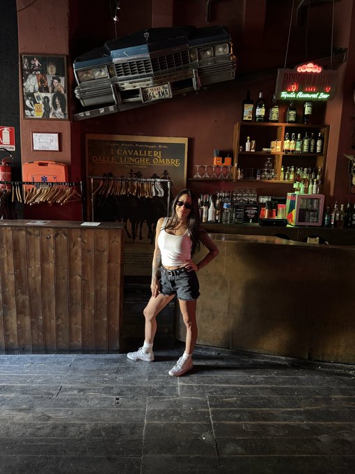 A woman poses in front of a vintage American car mounted above a bar, with visible signage reading "Cavalieri della Lunghe Ombre" and a Guinness display, capturing a quirky Italian spot during her tour.