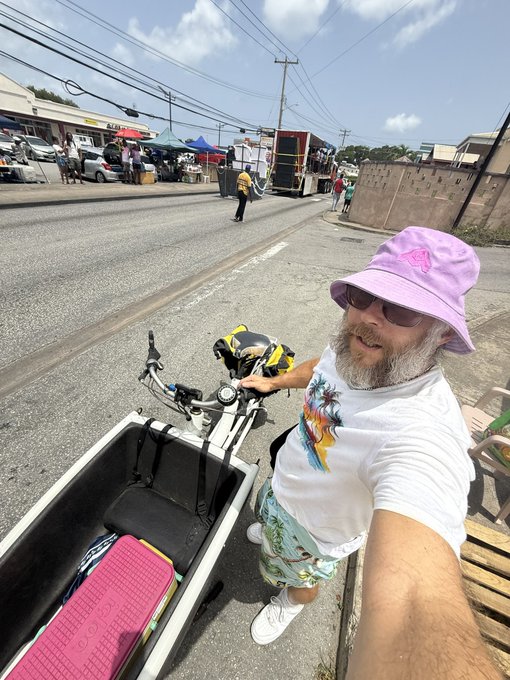 A selfie of a bearded man wearing a purple bucket hat and sunglasses, standing next to a cargo bike on a street, with a market visible in the background.