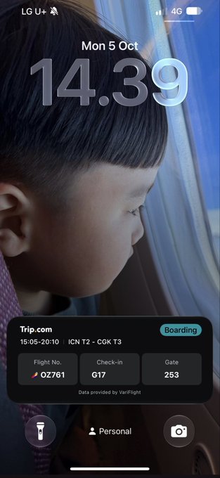An iPhone lock screen on LG U+ carrier showing the time 14:39 on Mon 5 Oct, with a Trip.com boarding pass widget displaying flight OZ761, check-in G17, gate 253 from ICN T2 to CGK T3 (15:05-20:10), over a photo of a child looking out a window.