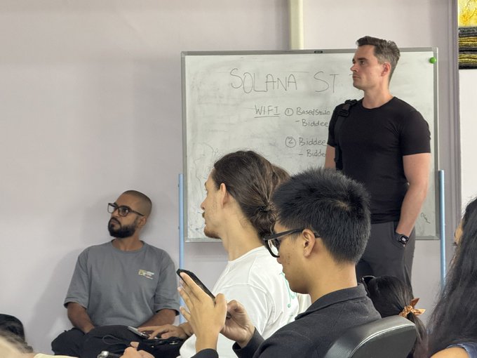 A classroom setting where a man in a black t-shirt stands by a whiteboard labeled 'SOLANA ST... WIFI 1/2' with notes about 'BaseStation' and 'Bidder', while students sit listening.