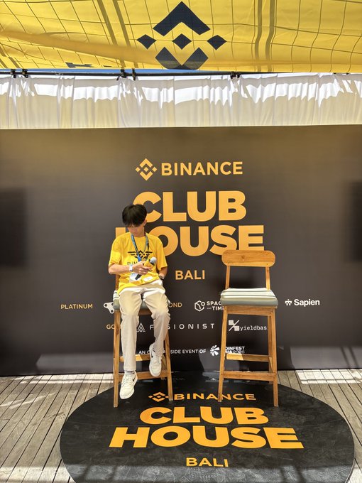 A person in a yellow Binance t-shirt sits on a stool looking at their phone at the Binance Clubhouse in Bali, with the venue's branded backdrop and floor mat clearly displaying "BINANCE CLUB HOUSE BALI" alongside sponsor logos like Platinum, Sapien, and Yieldbasis.