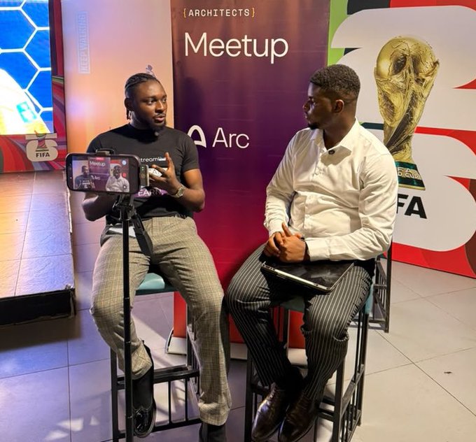 Two men are seated on stools during an "Architects Meetup" event, with one being filmed on a smartphone while speaking, against a backdrop featuring FIFA World Cup branding and Arc (Arc browser) sponsorship. The scene captures a live interview or panel discussion promoting Arc to an engaged audience.