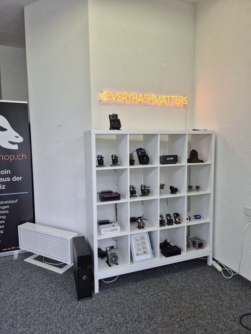 A display shelf filled with various mining rigs and electronic devices, topped with a neon sign that reads '#EVERYHASHMATTERS' and flanked by a banner for 'shop.ch'.