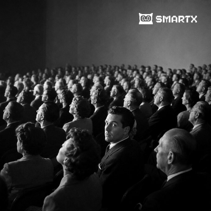A black and white photograph of an audience in a theater, with one man in the center turning to look at the camera, and a '0.0 SMARTX' watermark in the upper right corner.