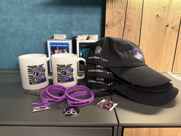 A display of merchandise including two white mugs with blue and yellow logos, two black baseball caps, and purple silicone wristbands arranged on a wooden surface.