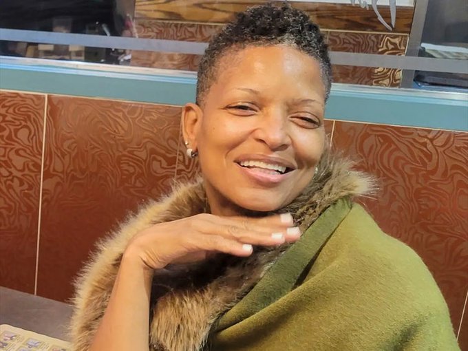 A smiling woman with short hair poses with her hand near her chin, wearing an olive green top with a fur-trimmed collar, in what appears to be a restaurant setting. The image accompanies the tragic news story about the missing New York City grandmother found dead near the Las Vegas airport after a stranger allegedly used her phone to request bitcoin.