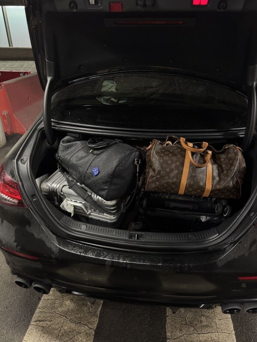 The open trunk of a black car reveals luggage inside, including a Louis Vuitton monogram duffle bag with tan leather handles and a dark backpack.
