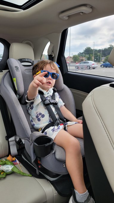 A toddler wearing sunglasses and a Mickey Mouse shirt sits in a Graco car seat, pointing toward the camera.