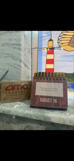An AMC Theatres business card and a wooden flip calendar dated August 16 sit on a counter, with a quote reading 'What comes easy won't last; what lasts won't come easy.'