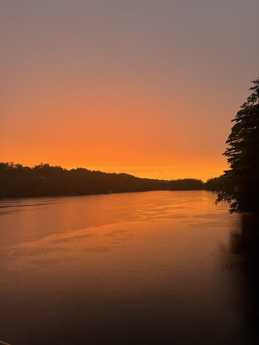 A serene sunset casts vibrant orange and amber hues across a calm lake, with silhouetted tree-lined shores framing the water. The sky transitions from deep orange at the horizon to soft lavender above, reflected perfectly on the glassy surface below.
