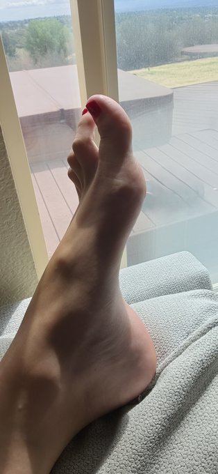 A close-up photo shows a person's bare foot resting on a textured white surface, featuring a red-painted toenail with a scenic landscape visible through a window in the background.
