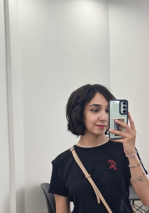 A woman with short dark hair takes a mirror selfie using a mint green phone case, wearing a black t-shirt with a small red logo and a beige crossbody strap.