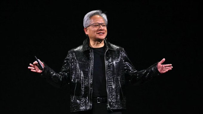 A man with gray hair and glasses wearing a black crocodile-patterned leather jacket, standing on a dark stage with arms outstretched while holding a small device.