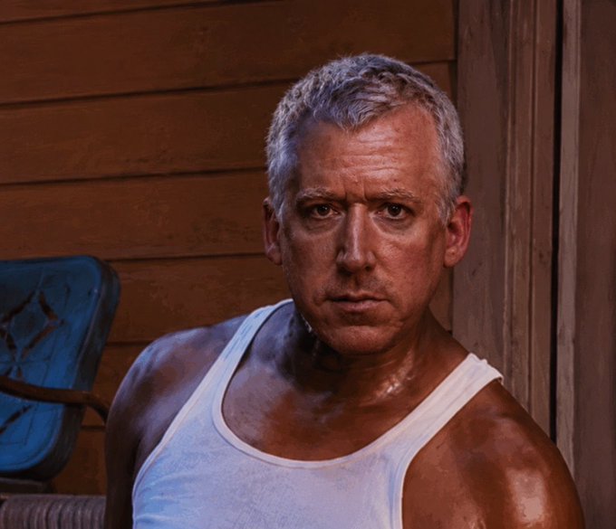 A muscular man with short gray hair wearing a white tank top, sitting outdoors near a wooden building with a serious expression.