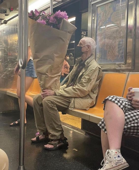 An elderly man wearing a face mask sits on a New York City subway train (42 St station visible), holding a massive bouquet of purple flowers wrapped in brown paper.