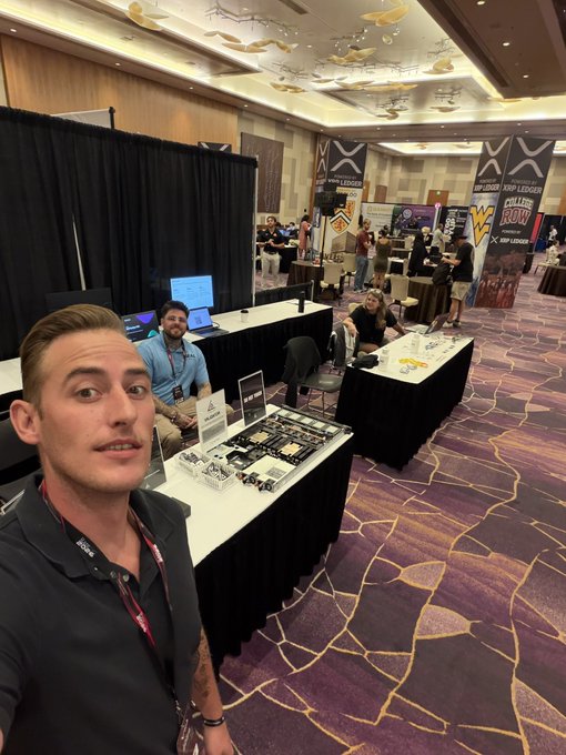 A man takes a selfie at a busy convention hall featuring an "XRPL LEDGER" and "JOIN LEDGER" banner in the background, with a table displaying computer hardware components nearby.