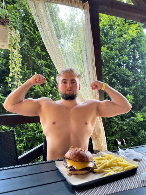 A shirtless 22-year-old flexes his biceps at an outdoor patio table featuring a cheeseburger and fries, seemingly celebrating his self-made success in crypto as referenced in the accompanying post about starting with just $15 from lunch money.