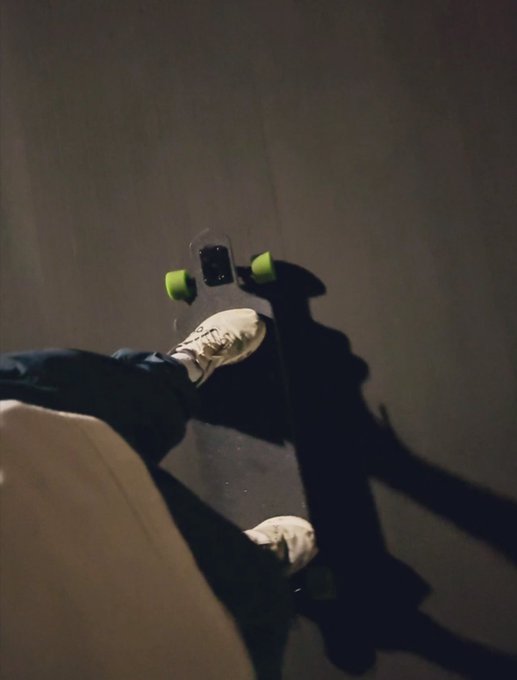 A person rides a longboard down a dark concrete ramp, their white sneakers visible as a strong light casts a dramatic shadow of them and the board against the wall.