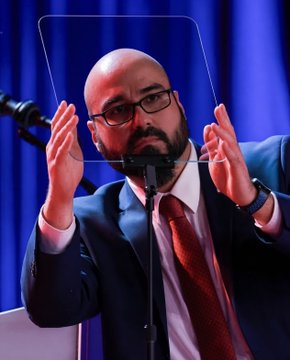 A bald man with glasses in a suit and red tie removing a teleprompter glass at a podium.