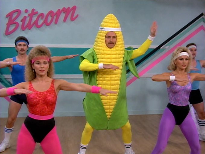 A still from an '80s-style workout video showing people in spandex aerobics gear doing exercises, with the central man wearing a full corn cob costume; the word 'Bitcorn' appears in pink retro text at the top.
