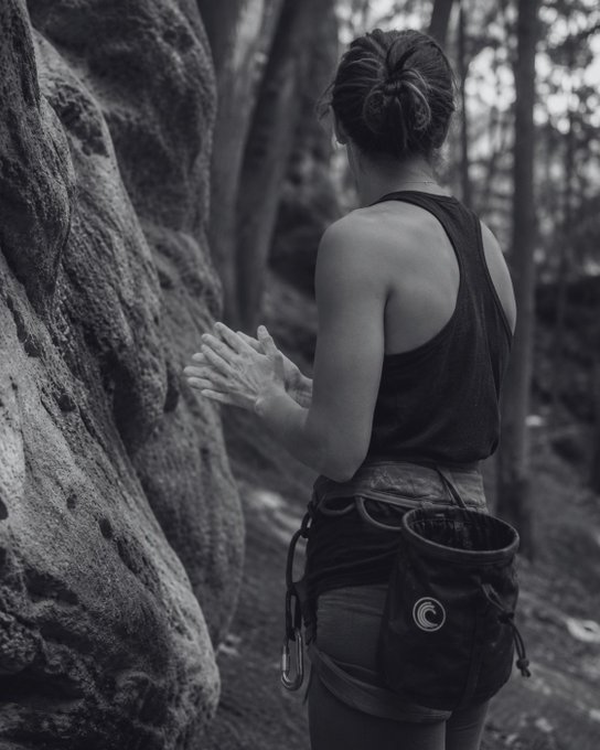 A woman with a chalk bag stands facing a boulder in a forest, rubbing chalk on her hands in preparation for rock climbing—a fitting visual for "GM. Powder up."