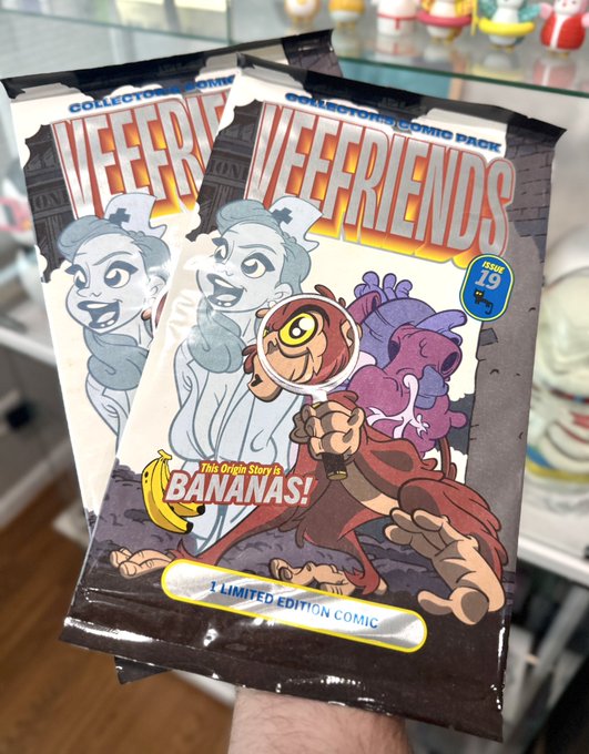 A hand holds a sealed 'VEEFRIENDS' collector's comic pack, Issue 19, featuring the character 'BANANAS' and labelled as '1 LIMITED EDITION COMIC' in a glass display case.