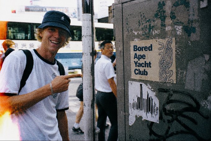 A person smiling and pointing at a 'Bored Ape Yacht Club' poster with a barcode design affixed to a street utility box.