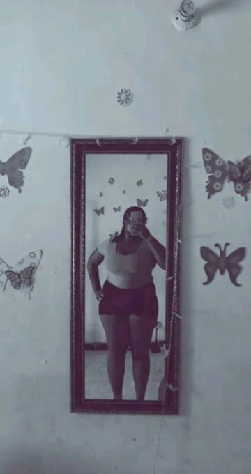 A person takes a mirror selfie in a tall framed mirror hanging on a white wall decorated with butterfly decals, wearing a crop top and dark shorts.
