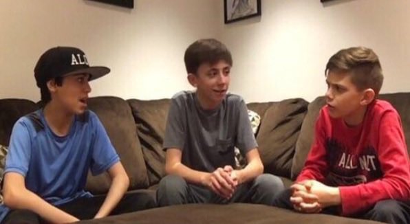 Three boys sit on a couch having a conversation, with one wearing a black cap that says "ALL" and another wearing a red "ALL DAY" hoodie.