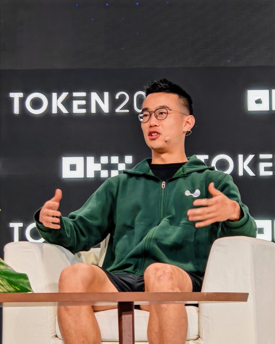 A man wearing glasses and a green hoodie sitting on a white chair on stage at the TOKEN 2049 event, gesturing with his hands while speaking.