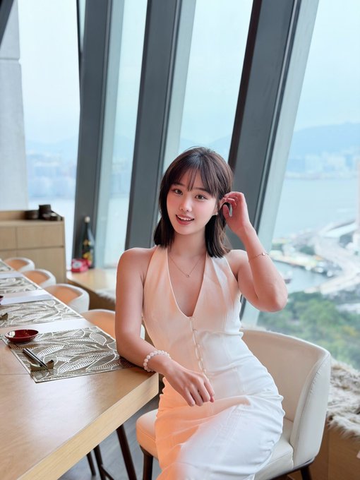 The image features a woman sitting at a dining table with her legs crossed while wearing a white dress. She has her hands resting on her lap, giving off an elegant and relaxed vibe. The background of the photo shows a window overlooking water, adding a serene atmosphere to the scene. There are chairs around the dining table, suggesting that it is likely a restaurant or a similar setting where people gather for meals.