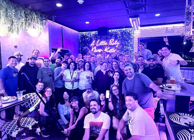 A large group of people pose together for a group photo in a purple-lit venue with neon signage that reads 'A Little Poetry Never Killed Nobody,' with many attendees smiling and raising their hands.