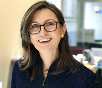 A smiling woman with long brown hair and black-rimmed glasses, wearing a dark blue blazer and gold earrings, is shown in what appears to be a professional office setting. The image represents Cathie Wood, whose Ark Invest firm made a significant purchase of Circle Internet Group ($CRCL) shares amid the stock's recent decline.