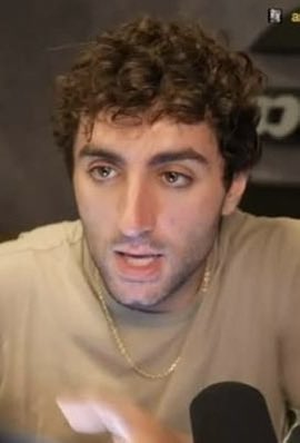 A young man with curly brown hair and a gold chain is shown speaking into a microphone, wearing a beige t-shirt. His intense, focused expression suggests he is reacting to or discussing the breaking news about $CATE's surge in market cap contrasted with RASMR's decline.