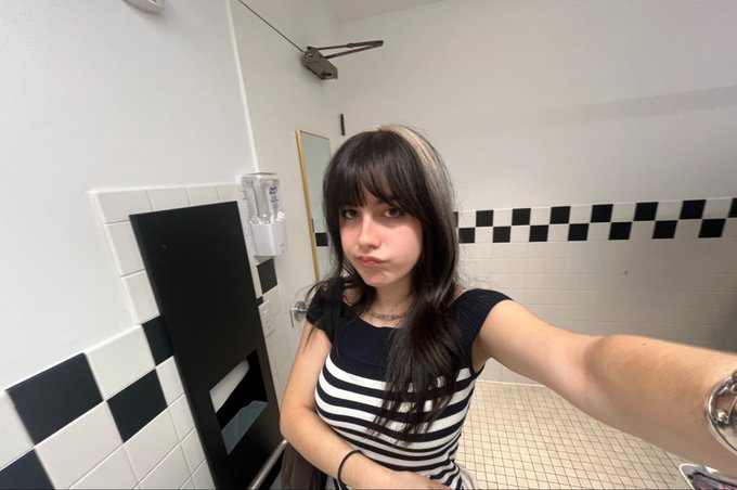 A young woman with long dark hair and bangs takes a mirror selfie in a black-and-white tiled bathroom, wearing a striped top and a silver watch while making a subtle pout. The image relates to the post's humorous observation that she resembles the iconic AI researcher associated with Nous Research.