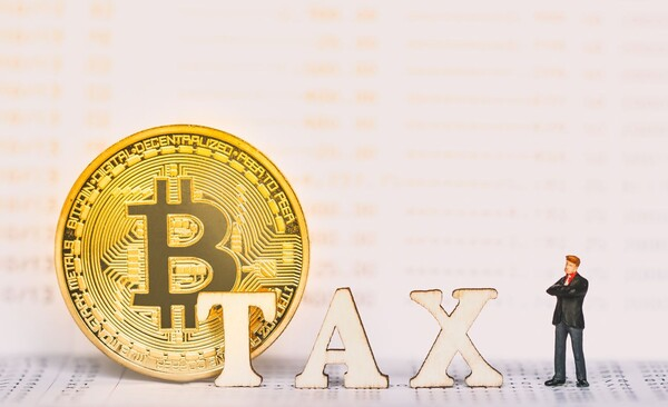 A golden Bitcoin coin sits beside the word "TAX" spelled out in wooden letters, with a miniature figurine of a man in a suit standing next to it, illustrating the ongoing legislative debate over cryptocurrency taxation in South Korea.