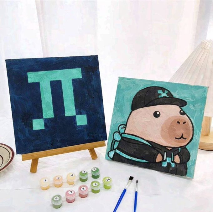 The image shows a table with two paintings, one depicting a cartoon pig wearing a black hat and the other featuring a blue square with a green triangle inside. The paintings are displayed on easels next to each other. There are also paintbrushes and paint tubes scattered around the table, suggesting that someone is working on an art project or preparing for a painting session.