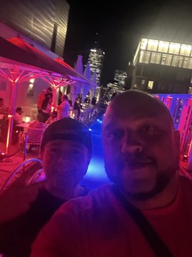 A nighttime selfie of a man and woman at an outdoor venue illuminated with vibrant pink and blue neon lights, with city buildings visible in the background.