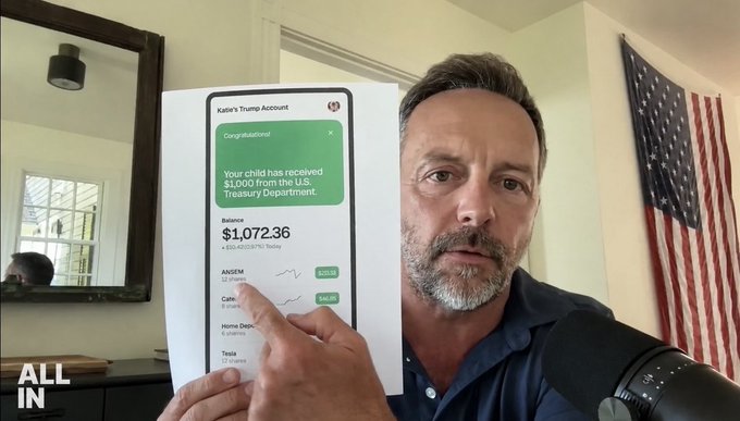 A man with gray hair and a beard holds up a printout of a Trump account showing a balance of $1,072.36, with an 'ALL IN' logo in the corner and an American flag in the background.