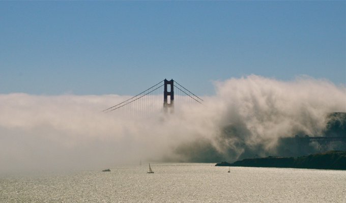 The Golden Gate Bridge in San Francisco emerges mysteriously from a thick blanket of fog rolling in over the bay, its iconic red tower partially obscured by the swirling mist. This atmospheric scene perfectly captures the challenge theme, showcasing nature's dramatic interplay between the famous landmark and the dense coastal fog that often envelops the bridge.