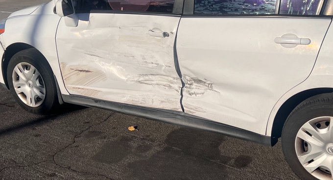 A white SUV with significant scrape damage and dents along its side doors, parked on asphalt.