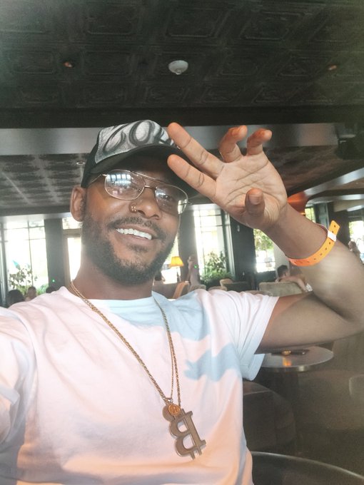 A smiling man wearing glasses, a white t-shirt, a Bitcoin pendant necklace, and a cap takes a selfie while raising his hand in a gesture, with an orange wristband visible on his wrist.