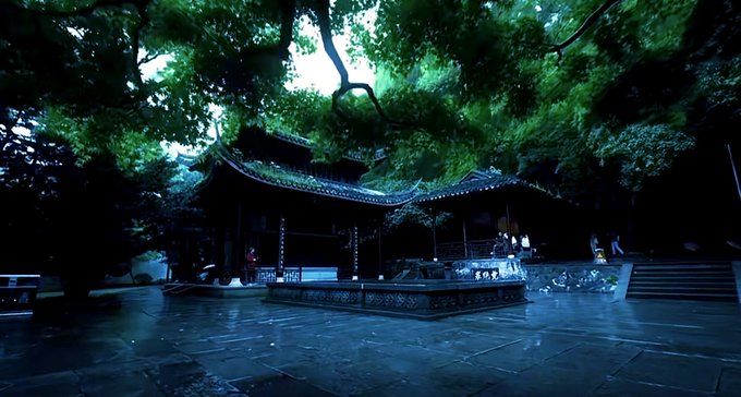 A serene, dimly-lit courtyard featuring traditional East Asian architecture, surrounded by lush green trees and stone paving, with a few visitors visible near the structure.
