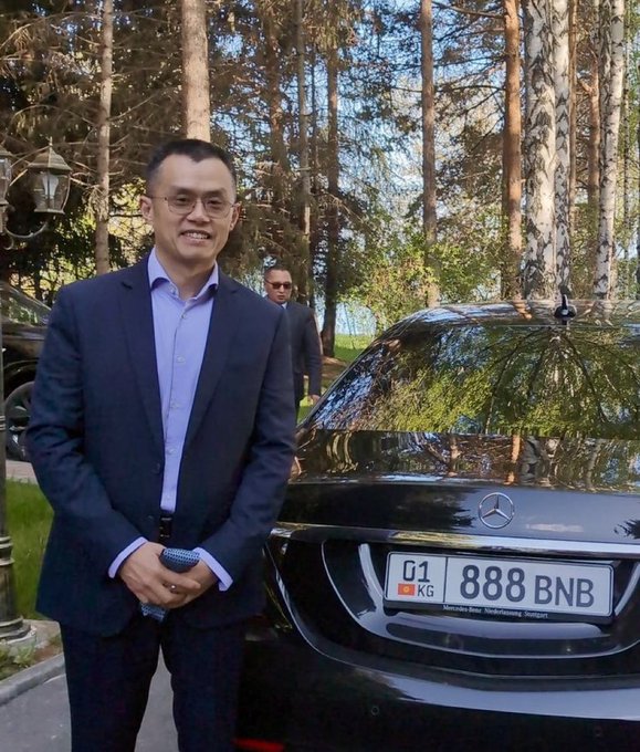 A smiling man in a dark suit stands beside a black Mercedes with the license plate "888 BNB," seemingly referencing the BNB cryptocurrency ticker and the auspicious number 888. The setting appears to be an upscale outdoor location with birch trees in the background, and a man in sunglasses stands behind near the vehicle.